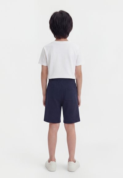 Παιδί που στέκεται με την πλάτη, φοράει λευκό T-shirt, σκούρα μπλε σορτς με πίσω τσέπη και λευκά αθλητικά παπούτσια σε απλό φόντο.