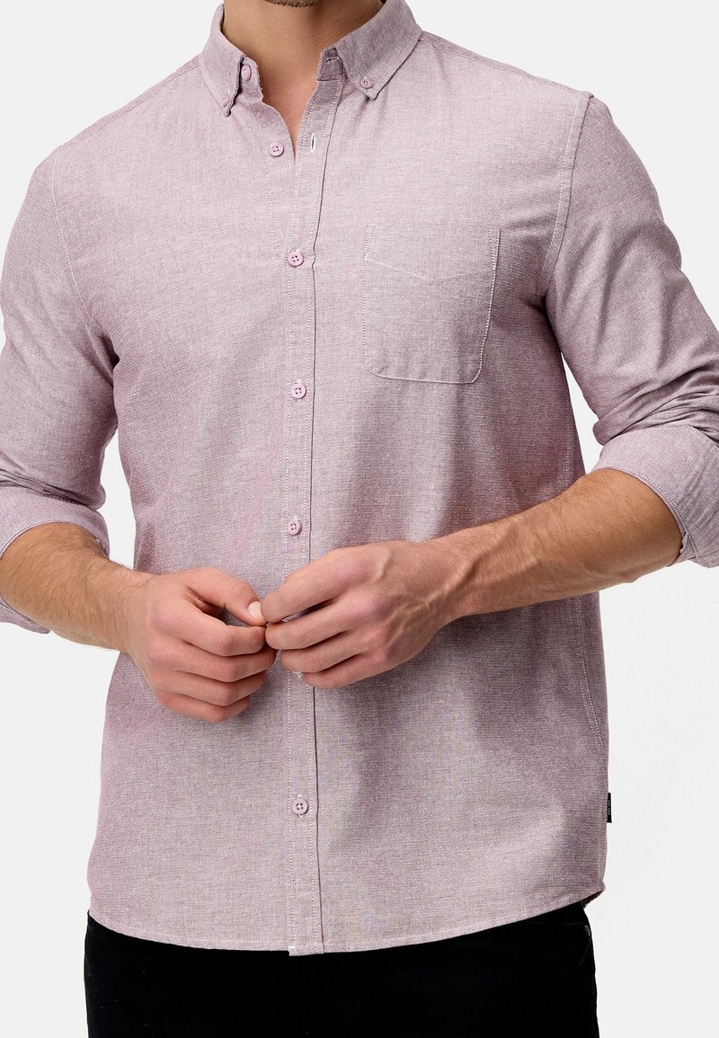 Homme portant une chemise rose pâle à manches longues avec manches retroussées, boutonnant les boutons du milieu, poche poitrine visible sur le côté gauche.
