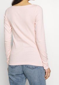 Langärmliges, hellrosa Oberteil mit Boot-Ausschnitt, aus weichem Stoff mit taillierter Silhouette, getragen zu blauen Jeans.
