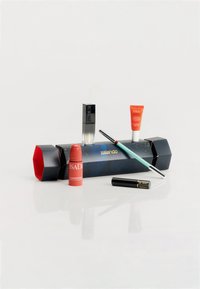 Prodotti cosmetici su una superficie riflettente bianca: un mascara nero, un blush rosso in stick, un eyeliner verde acqua e una crema idratante con vari packaging.