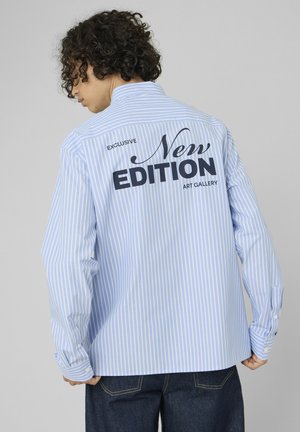 Personne aux cheveux bouclés portant une chemise rayée bleu et blanc avec le texte "Exclusive New Edition Art Gallery" dans le dos.