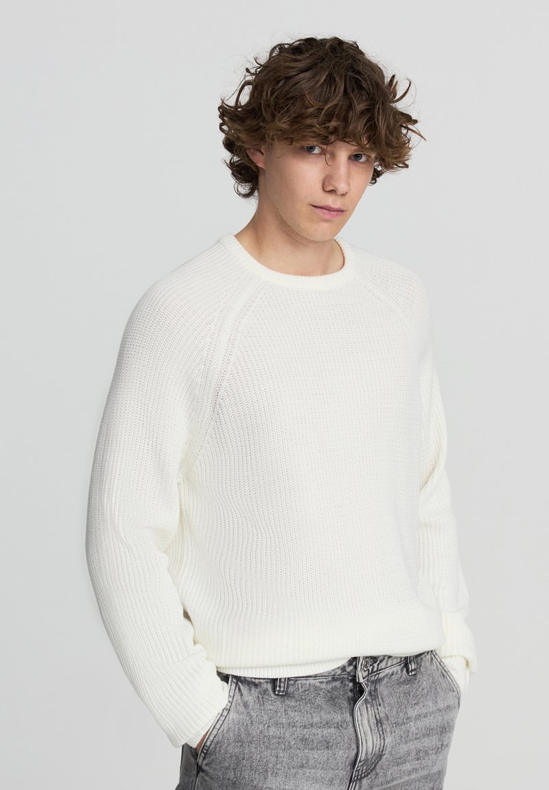 Biały, strukturalny sweter z okrągłym dekoltem i rękawami raglanowymi, w zestawieniu z jasnoszarymi dżinsami.