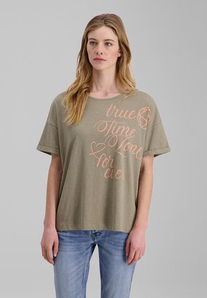 Young woman with long blonde hair wearing an olive green T-shirt with pink text "true Time Zone for ever" and blue jeans against a gray background.