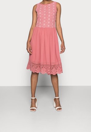 Vrouw draagt een mouwloze roze jurk tot op de knie met een kanten lijfje en zoom, gecombineerd met witte sandalen met hoge hakken, staand op een grijze vloer.