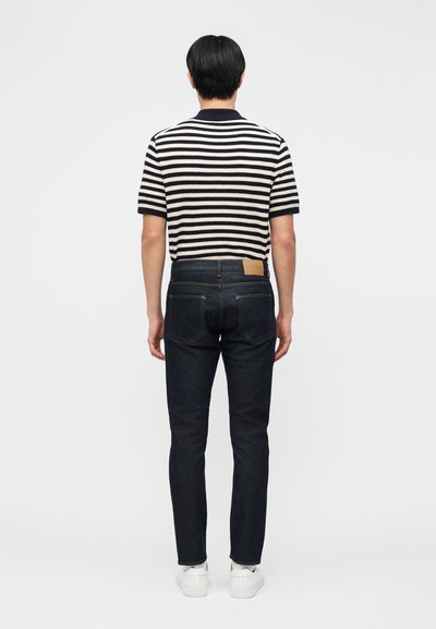 Muž v černo-bílém pruhovaném krátkém tričku, tmavě modrých džínách a bílých teniskách, stojící zády k nám na jednobarevném pozadí.