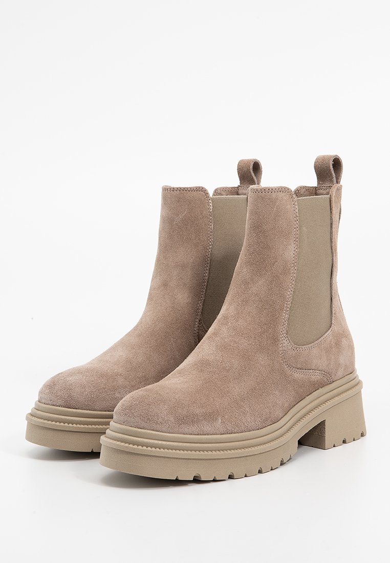 Para beżowych zamszowych botków Chelsea z elastycznymi panelami bocznymi, pętelkami do zakładania i masywnymi gumowymi podeszwami na białym tle.