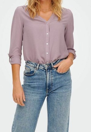 Blusa a pulsante di colore viola chiaro, con scollo a V e maniche arrotolate. Abbinata a jeans blu a vita alta, con una classica texture in denim.