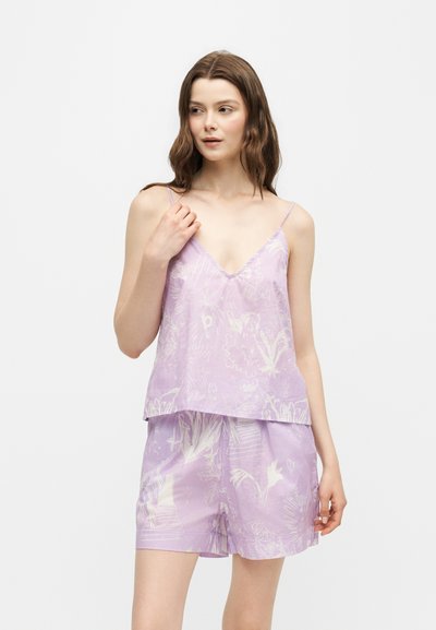Jeune femme portant un haut sans manches floral violet clair et un short assorti, debout devant un fond blanc uni.