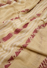 Beżowy materiał lniany z czerwonymi wzorami geometrycznymi i białymi poziomymi paskami, prezentujący miękką, teksturowaną powierzchnię i naturalny wygląd.