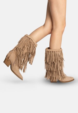 Piernas con botas de ante color canela hasta la mitad de la pantorrilla, con flecos en capas y tacones de madera apilados, mostradas sobre un fondo blanco liso.