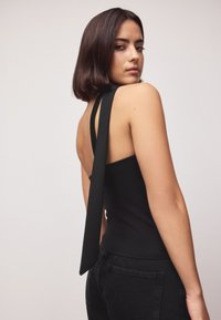 Femme aux cheveux courts et foncés portant un haut noir sans manches avec une longue lanière nouée dans le dos, associée à un pantalon noir, sur un fond uni.