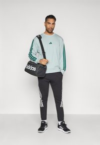 Ανοιχτό μπλε φούτερ με πράσινες ρίγες, μαύρο αθλητικό παντελόνι με λευκές ρίγες, μαύρη τσάντα ώμου με λευκό λογότυπο και μαύρα αθλητικά παπούτσια.