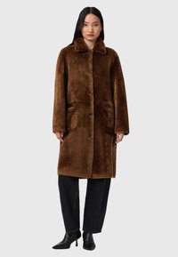 Cappotto in pelliccia sintetica marrone con colletto, chiusura a bottoni e due tasche frontali. Abbinato a jeans neri a vita alta e tacchi neri a punta.