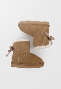Botas de invierno de ante marrón con forro de piel blanca y lazos de cinta marrón en la parte trasera, suelas de goma planas, mostradas desde vistas lateral y de la suela.