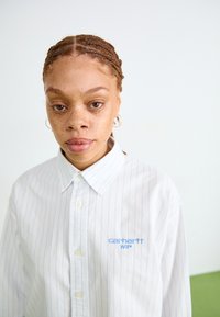 Camisa blanca de botones con rayas azul claro, un cuello puntiagudo y un logo de Carhartt WIP bordado en azul en el pecho.