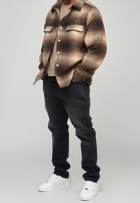 Rutig skjortjacka i brunt och beige med blå detaljer, med två bröstfickor och vita knappar. Svarta byxor och vita sneakers kompletterar outfiten.