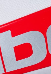Plastica rossa con scritte metalliche argento su uno sfondo bianco testurizzato; il design presenta caratteri audaci e un forte contrasto di colori.