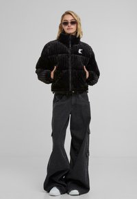 Black, ribbed puffer jacket with high collar and front zipper. Features white logo on chest, paired with baggy dark gray jeans and white sneakers.
