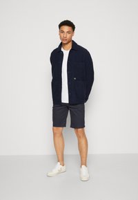 Marineblå kordfløyelsjakke med to frontlommer, hvit t-skjorte, mørke shorts og beige sneakers. Modellen står foran en ensfarget bakgrunn.