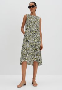 Blumenkleid mit grünem, schwarzem und weißem Muster, ärmelloses Design und fließendem Saum. Das Model trägt Sonnenbrillen und Riemchenschuhe.