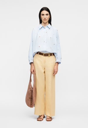 Woman with straight dark hair wears loose light blue embroidered shirt, beige wide-leg pants, brown belt, sandals, and holds a woven brown handbag.