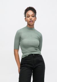 Φόρεμα polo σε ανοιχτό πράσινο με ριμπ ύφανση και μανίκια κοντά, συνδυασμένο με ψηλόμεσα μαύρα τζιν. Ομαλή υφή, εφαρμοστό σχέδιο.