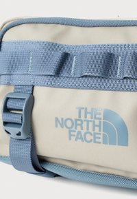 Μπεζ υφασμάτινη θήκη με μπλε λουριά και διακοσμητικά στοιχεία. Διαθέτει ένα εμφανές μπλε λογότυπο, ανθεκτική αγκράφα και υφή στην επιφάνεια. Συμπαγές ορθογώνιο σχήμα.