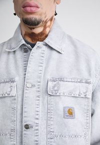 Gray denim jacket with two chest pockets, silver hardware, and a Carhartt logo patch. Features a buttoned collar and stitched detailing.