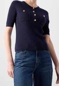 Granatowy prążkowany top z krótkim rękawem, z dwoma kieszeniami na piersi, złotymi guzikami i zaokrąglonym dekoltem. Połączony z niebieskimi dżinsami.