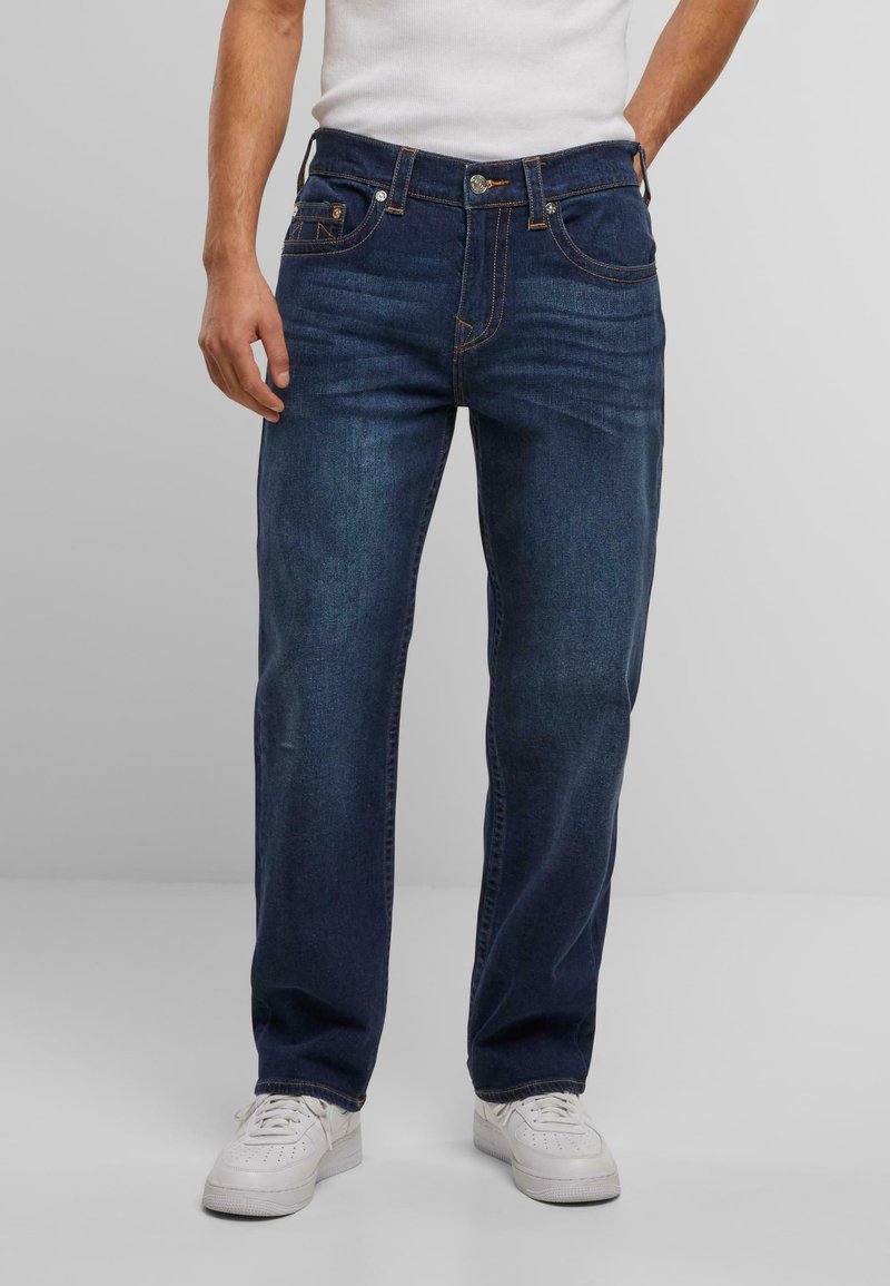 Jeans de mezclilla azul oscuro con corte recto, cinco bolsillos y detalles de costura naranja. Combinados con zapatillas blancas.