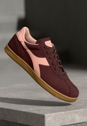 Maroon and pink sneaker with gum sole, detailed stitching, and laces, displayed mid-air above grey stone surface.