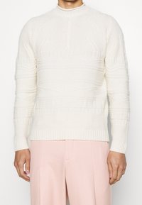 Pull en tricot crème avec des motifs texturés, col montant et poignets côtelés, associé à un pantalon tailleur rose clair.