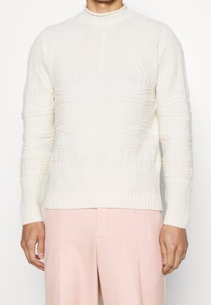 Cremfarbener Strickpullover mit strukturierten Mustern, hohem Kragen und gerippten Bündchen, kombiniert mit hellrosa maßgeschneiderten Hosen.