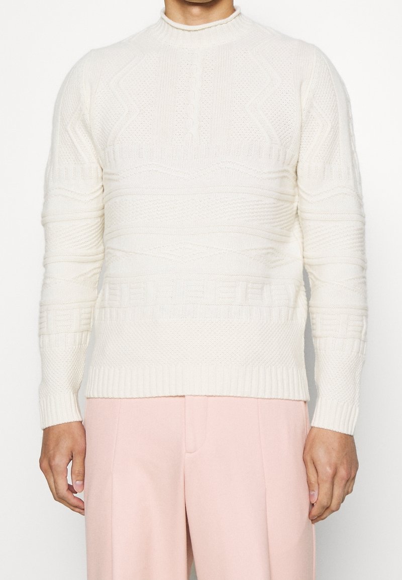 Pull en tricot crème avec des motifs texturés, col montant et poignets côtelés, associé à un pantalon tailleur rose clair.