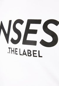 White t-shirt with large black text reading "SENSES .THE LABEL." Soft cotton material, rounded neckline, and short sleeves.