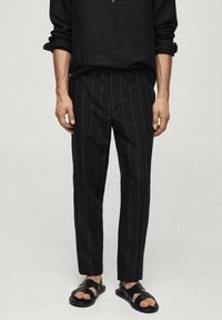 Camicia in lino nera con colletto abbottonato, abbinata a pantaloni neri a righe e sandali neri a fascia. Design semplice e dalla vestibilità rilassata.