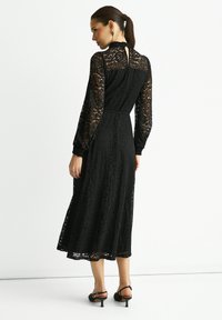 Black lace dress with long sheer sleeves, fitted waist, and flowing skirt. Features a high neckline and a keyhole back detail.