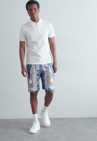 Weißes Polo-Shirt mit einem Reißverschlusskragen, blauen Shorts mit Blumenmuster und weißen Sneakers. Die Shorts verfügen über runde dekorative Elemente.