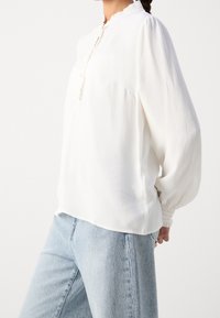 Kvinde iført en langærmet hvid struktureret bluse med volangkrave og lyseblå højtaljede jeans mod en ensfarvet baggrund.