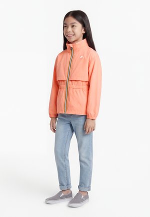 Νεαρή κοπέλα που φοράει κοραλλί ζακέτα με φερμουάρ, ανοιχτό μπλε τζιν με γυρισμένα μπατζάκια και γκρι παπούτσια χωρίς κορδόνια, στέκεται και χαμογελά.