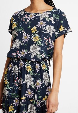 Vrouw draagt marineblauwe jurk met geel, wit en paars bloemenpatroon, korte mouwen en elastische taille, staand voor een witte achtergrond.