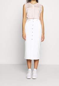 Jupe midi en denim blanc avec boutons sur le devant ; associée à un chemisier plissé rose clair avec bordure en dentelle et fermeture à boutons, sur un fond neutre.