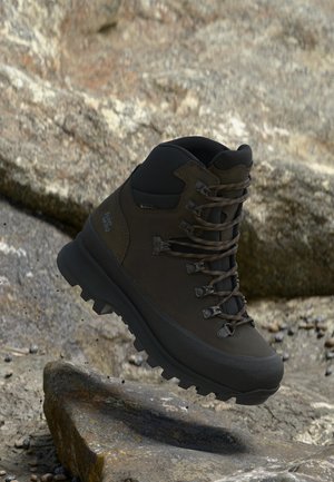 Bota de senderismo impermeable marrón con suela y cordones negros, suspendida sobre un terreno rocoso al aire libre.