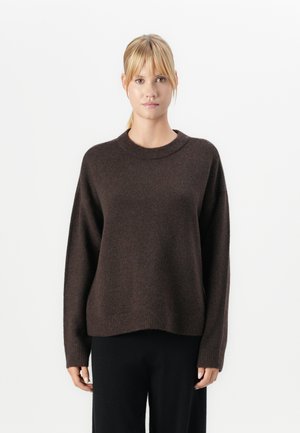Ung kvinna med blont hår, iklädd en mörkbrun stickad tröja och svarta byxor, stående mot en vit bakgrund.