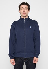 Felpa zip-up blu navale con colletto alto, realizzata in tessuto morbido. Presenta un piccolo logo circolare sul lato sinistro del petto e polsini e orlo a costine.