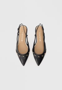 Zapatos planos slingback negros con punta puntiaguda, adornados con tachuelas decorativas doradas y plantillas color marrón claro con la marca GUESS, vistos desde arriba sobre un fondo blanco.