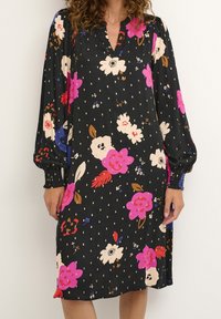 Kvinna bär en knälång svart klänning med rosa, vit, röd och blå blommönster och långa ärmar med smockade manschetter.