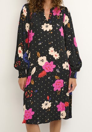 Una donna indossa un vestito nero a lunghezza ginocchio con una stampa floreale dai toni rosa, bianco, rosso e blu e maniche lunghe con polsini arricciati.