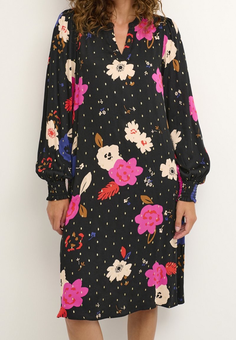Kvinna bär en knälång svart klänning med rosa, vit, röd och blå blommönster och långa ärmar med smockade manschetter.