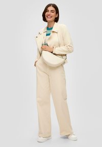 Cremfarbene Jacke mit Reißverschluss, gestreiftes Oberteil in Türkis und Weiß, cremfarbene weit geschnittene Hose und eine halbrunde Schultertasche. Weiße Sneakers.